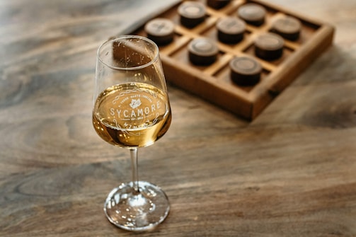 A glass of white wine with 'Sycamore' branding sits on a wooden surface. In the background, there is a wooden board game, possibly tic-tac-toe, with round game pieces.