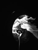 A close-up of swirling smoke illuminated against a dark background, creating a dramatic and mysterious effect. The smoke appears to be emanating from a small nozzle or tip, with intricate patterns and textures visible.