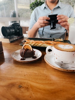 A person in a light blue shirt is holding a small camera, apparently taking a picture of food on the table. The wooden table has a plate with a pastry and a cup of coffee with latte art. Next to it, there's a chocolate dessert garnished with an orange slice. A large camera is visible on the left side of the image.