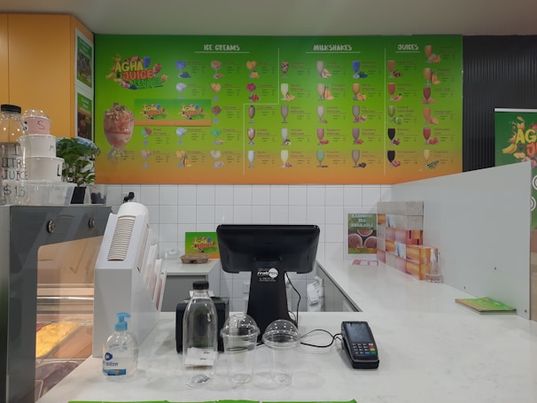 A juice bar counter with a vibrant menu displayed on the wall featuring various ice cream, milkshakes, and juice options. The countertop holds a cash register, payment terminal, sanitizer bottle, empty cups, and a water bottle. The color scheme consists of green and orange tones, with a partial view of product storage and promotional signage.