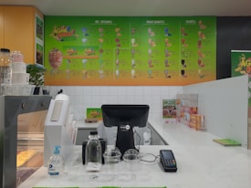 A juice bar counter with a vibrant menu displayed on the wall featuring various ice cream, milkshakes, and juice options. The countertop holds a cash register, payment terminal, sanitizer bottle, empty cups, and a water bottle. The color scheme consists of green and orange tones, with a partial view of product storage and promotional signage.