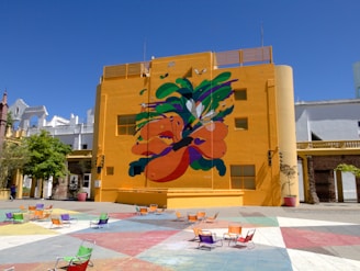 A vibrant mural with abstract shapes and organic forms covers a large part of a bright yellow building. The mural features a mix of bold colors such as orange, green, and purple. In front of the building, a plaza with multi-colored tiles and scattered colorful chairs can be seen. The sky is clear and blue, providing a striking contrast against the building.