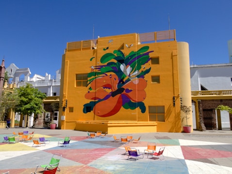 A vibrant mural with abstract shapes and organic forms covers a large part of a bright yellow building. The mural features a mix of bold colors such as orange, green, and purple. In front of the building, a plaza with multi-colored tiles and scattered colorful chairs can be seen. The sky is clear and blue, providing a striking contrast against the building.