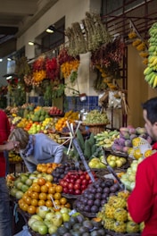 A bustling fruit market with a variety of colorful fruits displayed in baskets. Bananas, oranges, tomatoes, and other tropical fruits fill the scene. Hanging dried herbs and flowers add to the vibrant atmosphere. People are seen browsing and inspecting the produce.