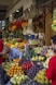 A bustling fruit market with a variety of colorful fruits displayed in baskets. Bananas, oranges, tomatoes, and other tropical fruits fill the scene. Hanging dried herbs and flowers add to the vibrant atmosphere. People are seen browsing and inspecting the produce.