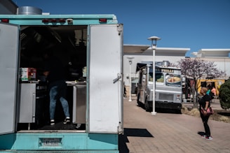 A scene featuring two food trucks parked in an outdoor area, with a person working inside the nearer blue food truck and a woman walking past on the right. A tree with purple blossoms is visible near the woman, and there are buildings in the background.
