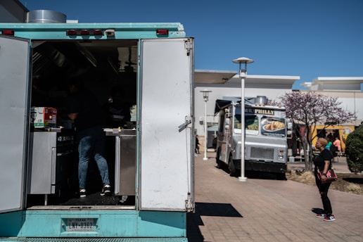 A scene featuring two food trucks parked in an outdoor area, with a person working inside the nearer blue food truck and a woman walking past on the right. A tree with purple blossoms is visible near the woman, and there are buildings in the background.