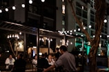 A bustling outdoor dining area is seen at night, featuring string lights that illuminate a group of people seated at tables. The ambiance is cozy and intimate, with warm-toned hanging bulbs adding to the atmosphere. A building with multiple floors and balconies provides a backdrop.