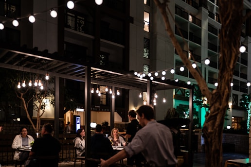A bustling outdoor dining area is seen at night, featuring string lights that illuminate a group of people seated at tables. The ambiance is cozy and intimate, with warm-toned hanging bulbs adding to the atmosphere. A building with multiple floors and balconies provides a backdrop.