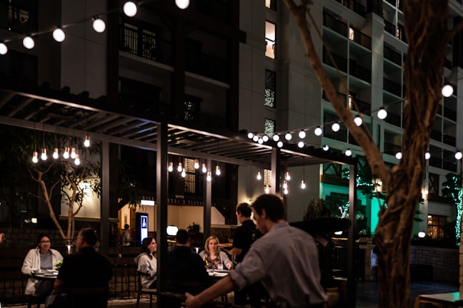 A bustling outdoor dining area is seen at night, featuring string lights that illuminate a group of people seated at tables. The ambiance is cozy and intimate, with warm-toned hanging bulbs adding to the atmosphere. A building with multiple floors and balconies provides a backdrop.