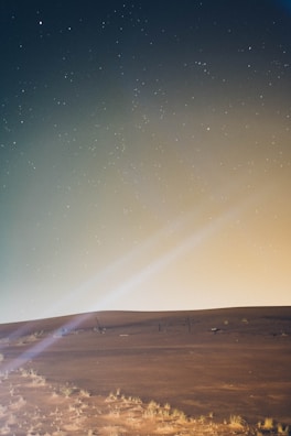 A serene landscape photo with beige sand dunes under a black night sky streaked with blue stars.
