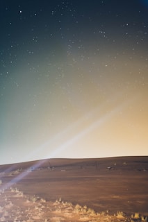 A serene desert landscape under a starry sky, with a gentle gradient from dark blue to a warm yellow at the horizon. The foreground shows sand dunes with sparse vegetation. Faint lines of starlight create a subtle, mystical atmosphere.