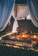 An intimate outdoor setting featuring a cozy arrangement of patterned pillows and rugs surrounded by small glowing candles. Sheer drapes hang in the background, creating an enclosed and inviting space. The scene is lit by soft lights under a night sky, suggesting a romantic or tranquil atmosphere.
