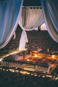 An intimate outdoor setting featuring a cozy arrangement of patterned pillows and rugs surrounded by small glowing candles. Sheer drapes hang in the background, creating an enclosed and inviting space. The scene is lit by soft lights under a night sky, suggesting a romantic or tranquil atmosphere.