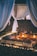 An intimate outdoor setting featuring a cozy arrangement of patterned pillows and rugs surrounded by small glowing candles. Sheer drapes hang in the background, creating an enclosed and inviting space. The scene is lit by soft lights under a night sky, suggesting a romantic or tranquil atmosphere.
