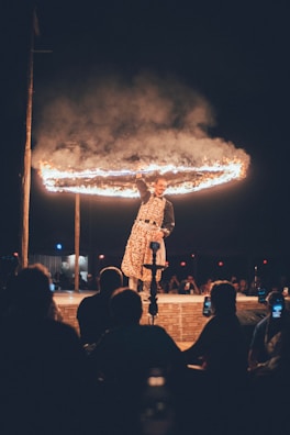 A performer dressed in patterned clothing stands on a stage holding a flaming hoop above their head. The background is dark, and a crowd of silhouetted spectators is visible seated in front of the stage, some using their phones to capture the moment.