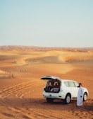 A white SUV is parked in a vast desert landscape with sandy dunes stretching into the distance. The vehicle's rear hatch is open, revealing some items inside. A person dressed in traditional white attire stands beside the SUV, gazing at the scenery. Tire tracks are visible in the sand, curving around the dunes.
