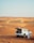 A white SUV is parked in a vast desert landscape with sandy dunes stretching into the distance. The vehicle's rear hatch is open, revealing some items inside. A person dressed in traditional white attire stands beside the SUV, gazing at the scenery. Tire tracks are visible in the sand, curving around the dunes.