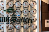 A wall is covered with rows of stainless steel kegs, each featuring a red circular logo in the center. In front of the kegs, there is a glass wall with the words 'Flying Saucer' in green, stylized lettering, accompanied by a UFO graphic.