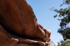 A large, smooth rock formation dominates the scene with its sweeping curves and varying shades of brown. A light fixture is attached to the rock, and the sky above is clear and bright. On the right, branches and leaves of a tree frame the image, adding a touch of green against the rock and blue sky.