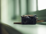 A digital camera sits on a glossy, soft-colored surface near a window. The camera is positioned at an angle that highlights its lens and metallic details. The lighting is gentle, casting subtle shadows and reflections on the surface, creating a calm and serene atmosphere.
