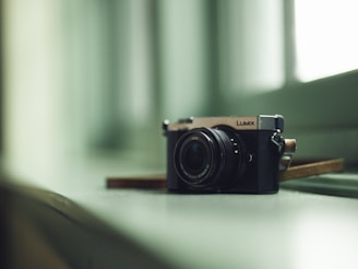 A digital camera sits on a glossy, soft-colored surface near a window. The camera is positioned at an angle that highlights its lens and metallic details. The lighting is gentle, casting subtle shadows and reflections on the surface, creating a calm and serene atmosphere.