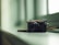 A digital camera sits on a glossy, soft-colored surface near a window. The camera is positioned at an angle that highlights its lens and metallic details. The lighting is gentle, casting subtle shadows and reflections on the surface, creating a calm and serene atmosphere.