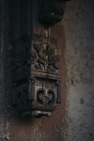 An intricately carved wooden structure with floral and geometric patterns is mounted on a textured wall. The wood appears aged, with visible cracks and a dark, rich patina.