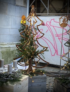 A metal art sculpture of a Christmas tree adorned with small string lights stands on a wooden base. Surrounding the sculpture are evergreen branches, and nearby sits a circular metal candleholder with four candles. The background includes a stone wall and a spray-painted canvas with abstract pink designs.