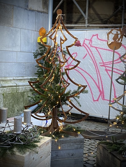 A metal art sculpture of a Christmas tree adorned with small string lights stands on a wooden base. Surrounding the sculpture are evergreen branches, and nearby sits a circular metal candleholder with four candles. The background includes a stone wall and a spray-painted canvas with abstract pink designs.