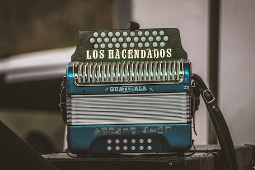 An accordion with the words 'LOS HACENDADOS' written on the keyboard section and 'GUATEMALA' on the body stands prominently. It features metallic buttons and a mix of blue and white colors. The background is slightly blurred, highlighting the instrument.
