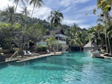 A serene tropical resort setting featuring a large swimming pool surrounded by lush greenery and palm trees. Lounge chairs are arranged on the poolside, and there are resort buildings with thatched roofs in the background. The sky is clear with few clouds, suggesting a sunny day, and the landscape includes a backdrop of forested hills.