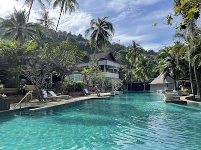 A serene tropical resort setting featuring a large swimming pool surrounded by lush greenery and palm trees. Lounge chairs are arranged on the poolside, and there are resort buildings with thatched roofs in the background. The sky is clear with few clouds, suggesting a sunny day, and the landscape includes a backdrop of forested hills.