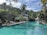 A serene tropical resort setting featuring a large swimming pool surrounded by lush greenery and palm trees. Lounge chairs are arranged on the poolside, and there are resort buildings with thatched roofs in the background. The sky is clear with few clouds, suggesting a sunny day, and the landscape includes a backdrop of forested hills.