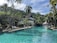 A serene tropical resort setting featuring a large swimming pool surrounded by lush greenery and palm trees. Lounge chairs are arranged on the poolside, and there are resort buildings with thatched roofs in the background. The sky is clear with few clouds, suggesting a sunny day, and the landscape includes a backdrop of forested hills.