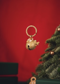 A golden fish-shaped bell is suspended in front of a festive red background. The bell is designed with intricate details and features a ring inscribed with the words 'GET BUSY LIVING OR GET BUSY DYING - COPPERISM'. To the right of the image, a portion of a decorated Christmas tree is visible, adorned with ornaments. In the lower part of the image, blurred elements suggest the presence of gift boxes.