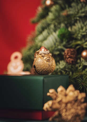 A gold ornament with intricate details sits atop a green gift box. In the background, a decorated Christmas tree with evergreen needles and small pinecones adds a festive touch. A blurred red background complements the scene.
