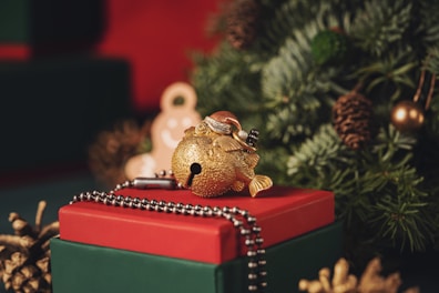 A close-up of a festive scene featuring a red and green gift box with a decorative golden fish ornament on top. The box is adorned with a beaded metal chain. In the background, there is a Christmas tree with pine cones and a blurred gingerbread figure.