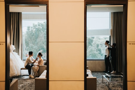 A scene divided by a wall, showing two separate rooms. On the left, a person is applying makeup to a seated bride, with a wedding dress on display nearby. The window provides a view of trees outside. On the right, a person is assisting a man in formal attire with adjusting his bow tie, also next to a window with a view of trees.