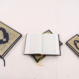 An open book with intricate Arabic script is positioned in the center, surrounded by two closed books with ornate gold detailing on their covers. One book is on the left and the other on the right, all placed on a light, neutral background.
