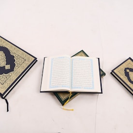An open book with intricate Arabic script is positioned in the center, surrounded by two closed books with ornate gold detailing on their covers. One book is on the left and the other on the right, all placed on a light, neutral background.