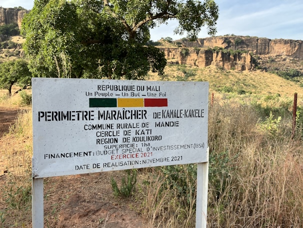 A rural sign in a grassy landscape with rocky cliffs in the background. The sign, written in French, references a market gardening area in the Kati region of Mali. Trees and foliage surround the area, under a clear sky.