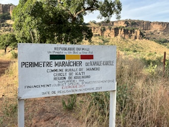 A rural sign in a grassy landscape with rocky cliffs in the background. The sign, written in French, references a market gardening area in the Kati region of Mali. Trees and foliage surround the area, under a clear sky.