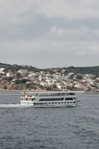 A large passenger ferry is moving across a body of water, with a backdrop of a hilly landscape filled with numerous white houses with red roofs. The houses appear densely packed, typical of a coastal town or village. The sky is overcast, contributing to a calm and serene atmosphere.