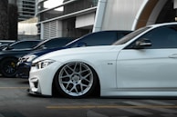 Several luxury cars are parked in a row in an urban setting with modern architecture in the background. The focus is on a sleek white car with custom wheels and low suspension.