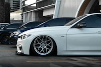 Several luxury cars are parked in a row in an urban setting with modern architecture in the background. The focus is on a sleek white car with custom wheels and low suspension.