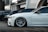 Several luxury cars are parked in a row in an urban setting with modern architecture in the background. The focus is on a sleek white car with custom wheels and low suspension.