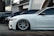 Several luxury cars are parked in a row in an urban setting with modern architecture in the background. The focus is on a sleek white car with custom wheels and low suspension.