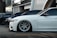 Several luxury cars are parked in a row in an urban setting with modern architecture in the background. The focus is on a sleek white car with custom wheels and low suspension.
