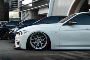 Several luxury cars are parked in a row in an urban setting with modern architecture in the background. The focus is on a sleek white car with custom wheels and low suspension.